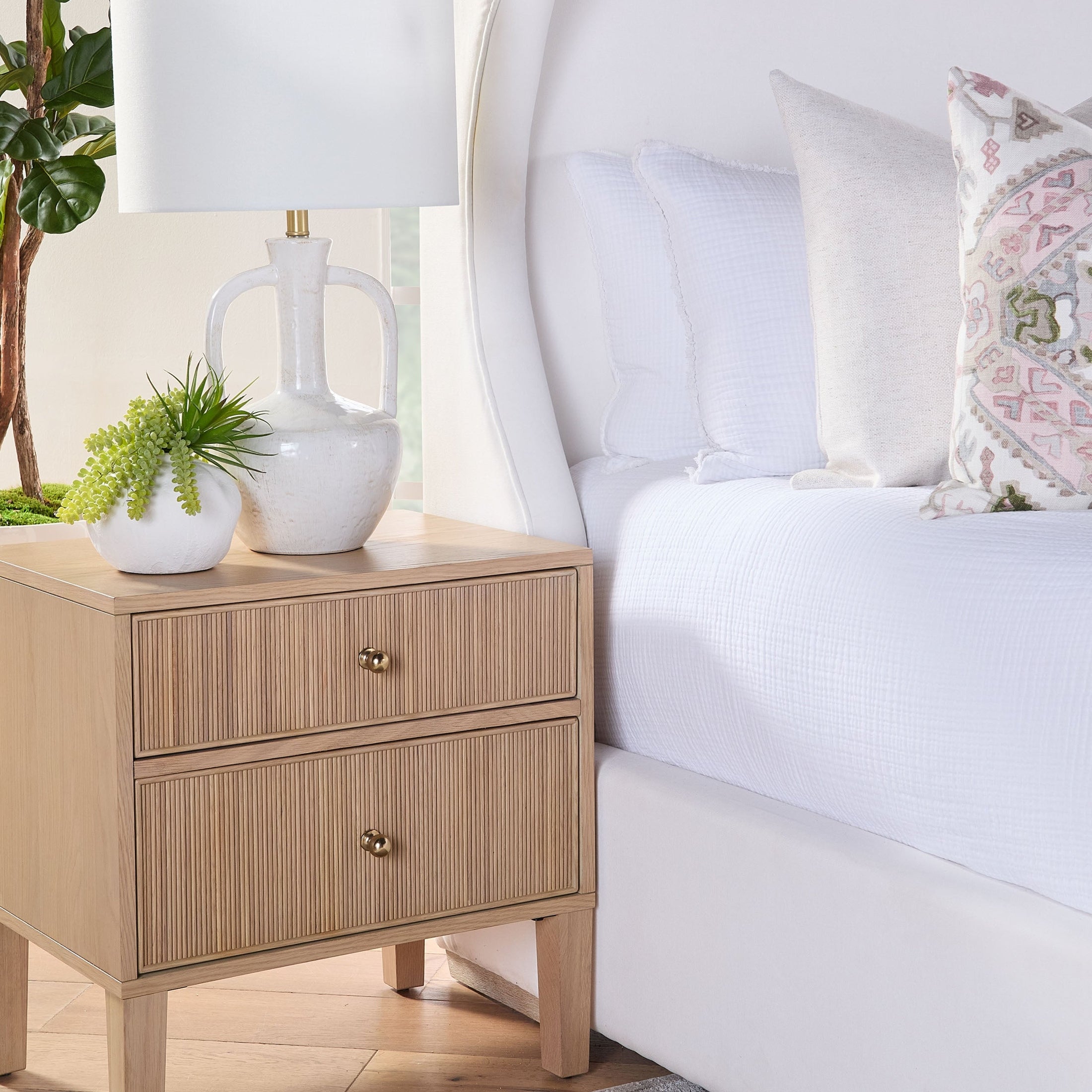 Highland 2-drawer nightstand in natural oak finish with brass knobs next to a white upholstered bed with pillows