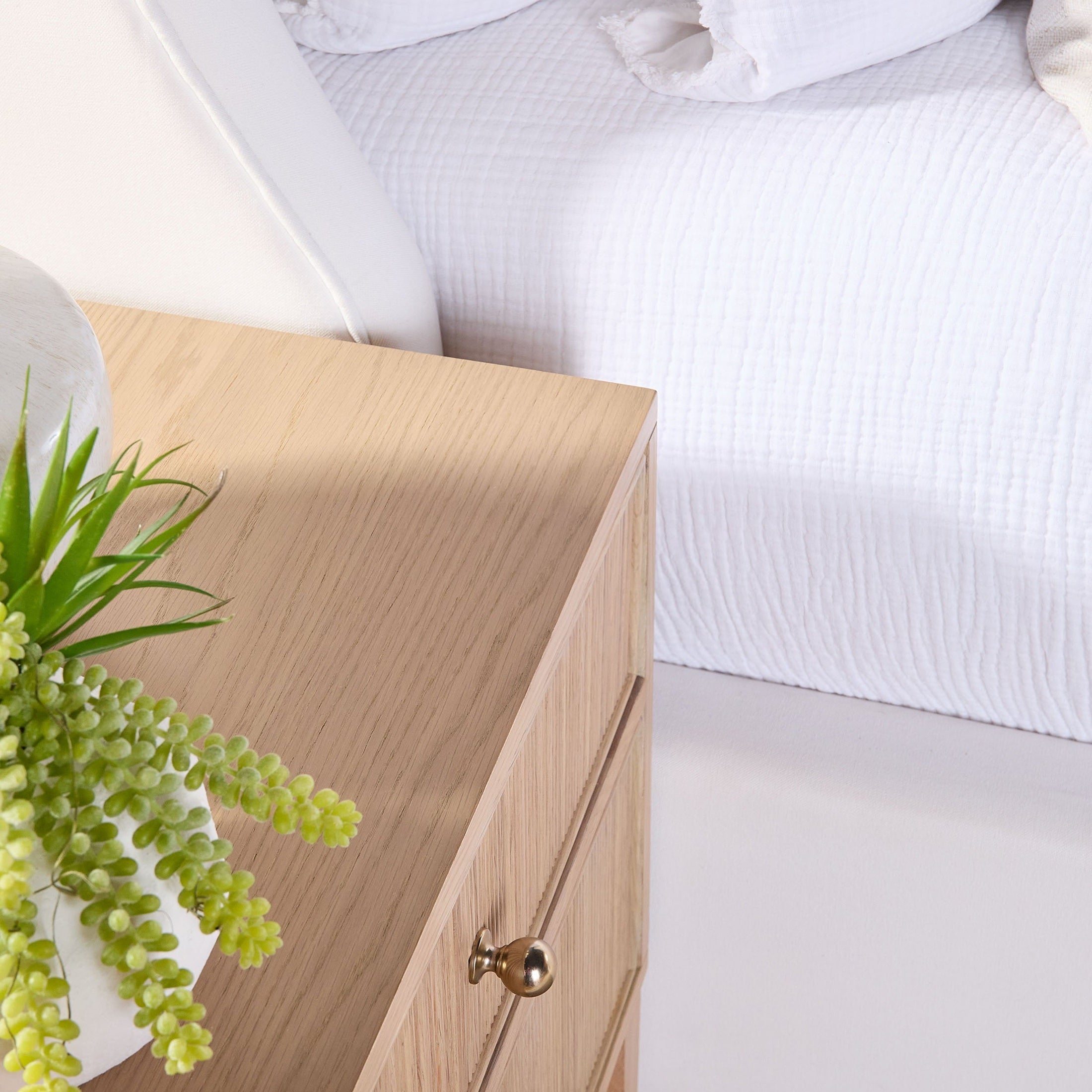 Close-up of Highland 2-Drawer Nightstand in natural oak finish with polished brass knob and textured drawer detail next to white bed