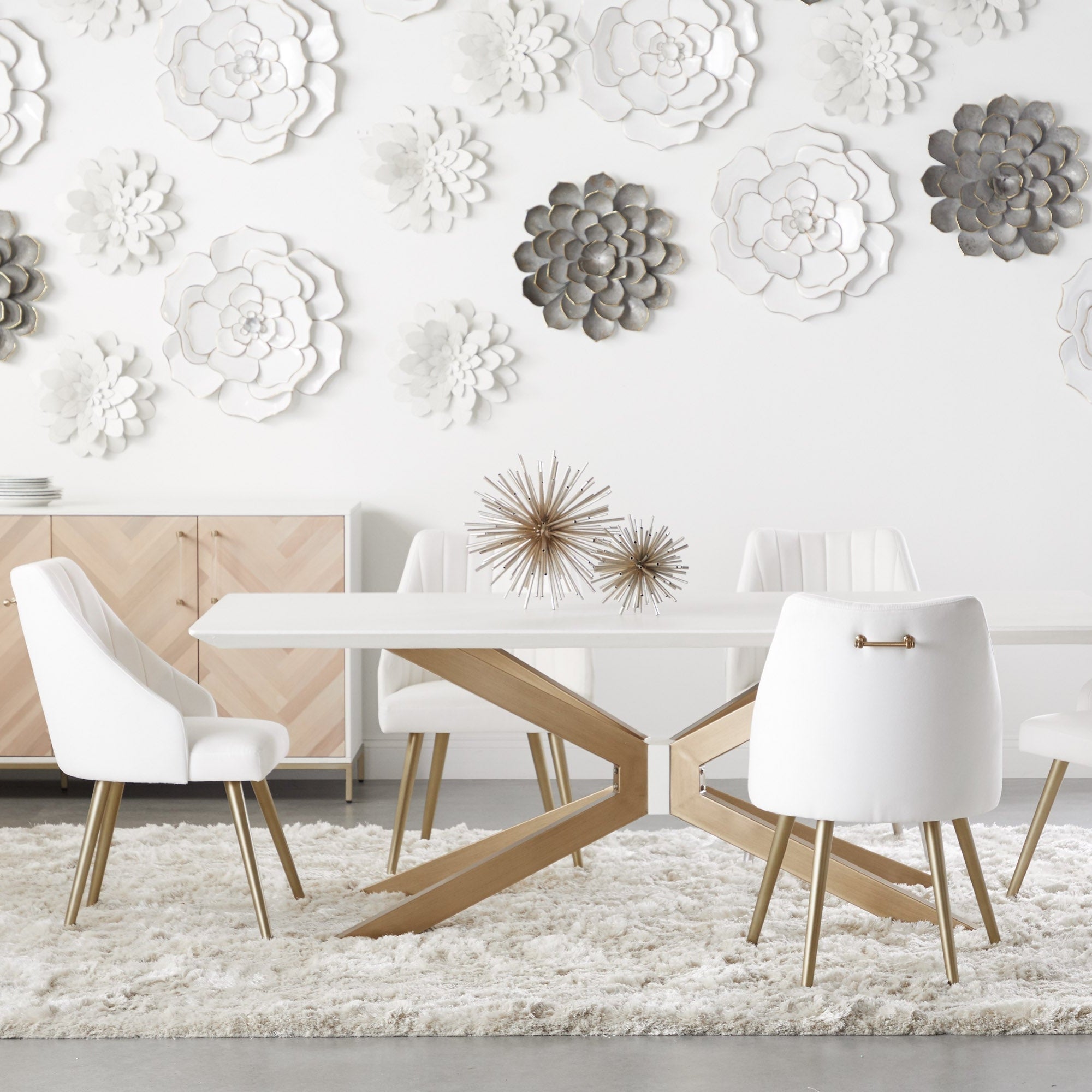 Modern white dining table with angled wood base and white upholstered chairs on plush rug in bright room