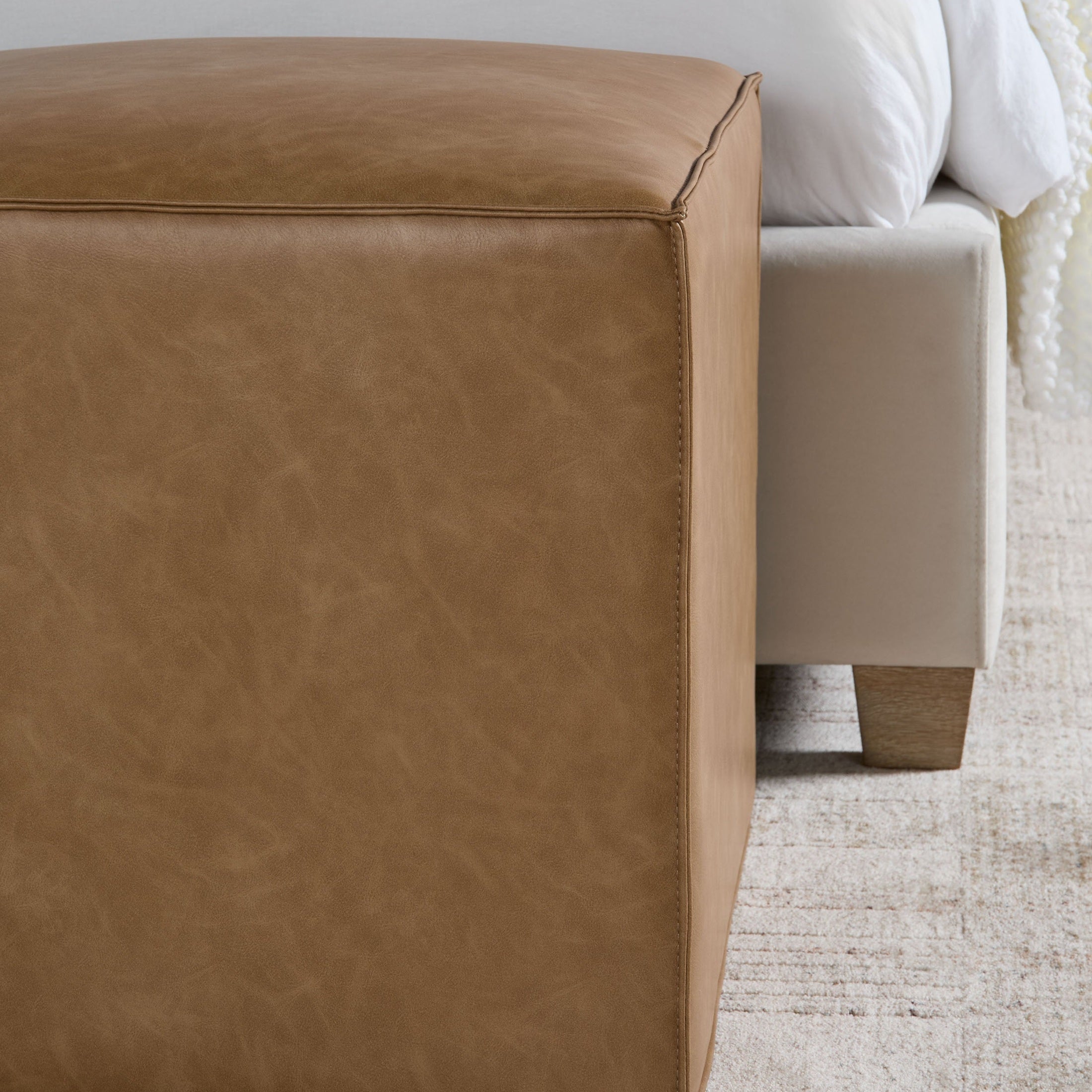 Knox Accent Cube by Essentials for Living in tan upholstery beside a bed on a textured rug in a modern room.