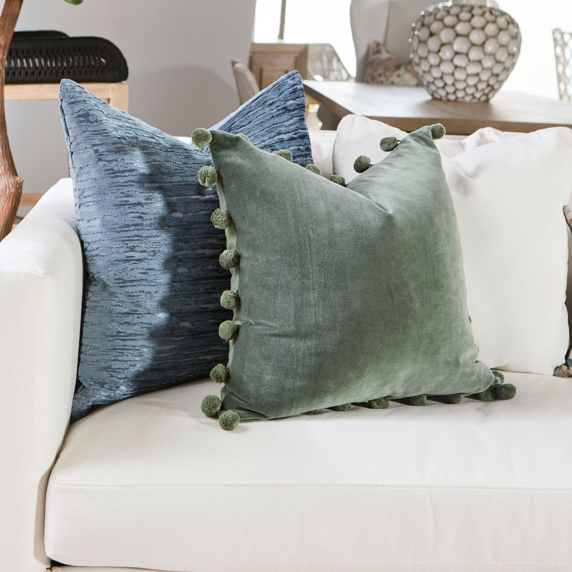 Two decorative velvet pillows in green and blue with pom-pom accents on a white sofa in a modern living room