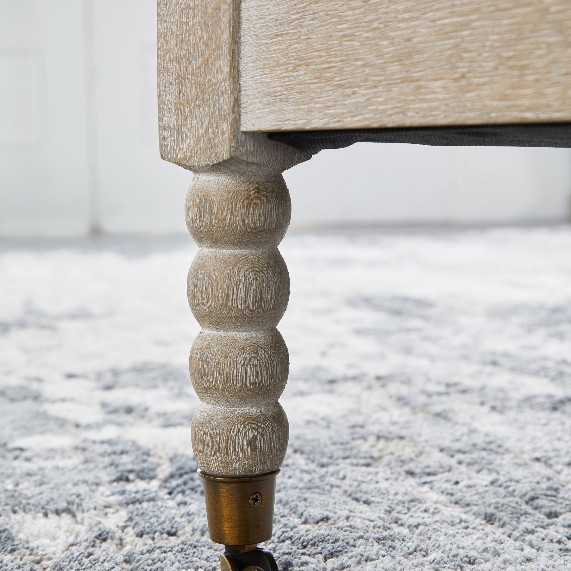 Close-up of the Rouleau Chaise Lounge natural gray oak carved spool-style leg with caster wheel on a textured rug