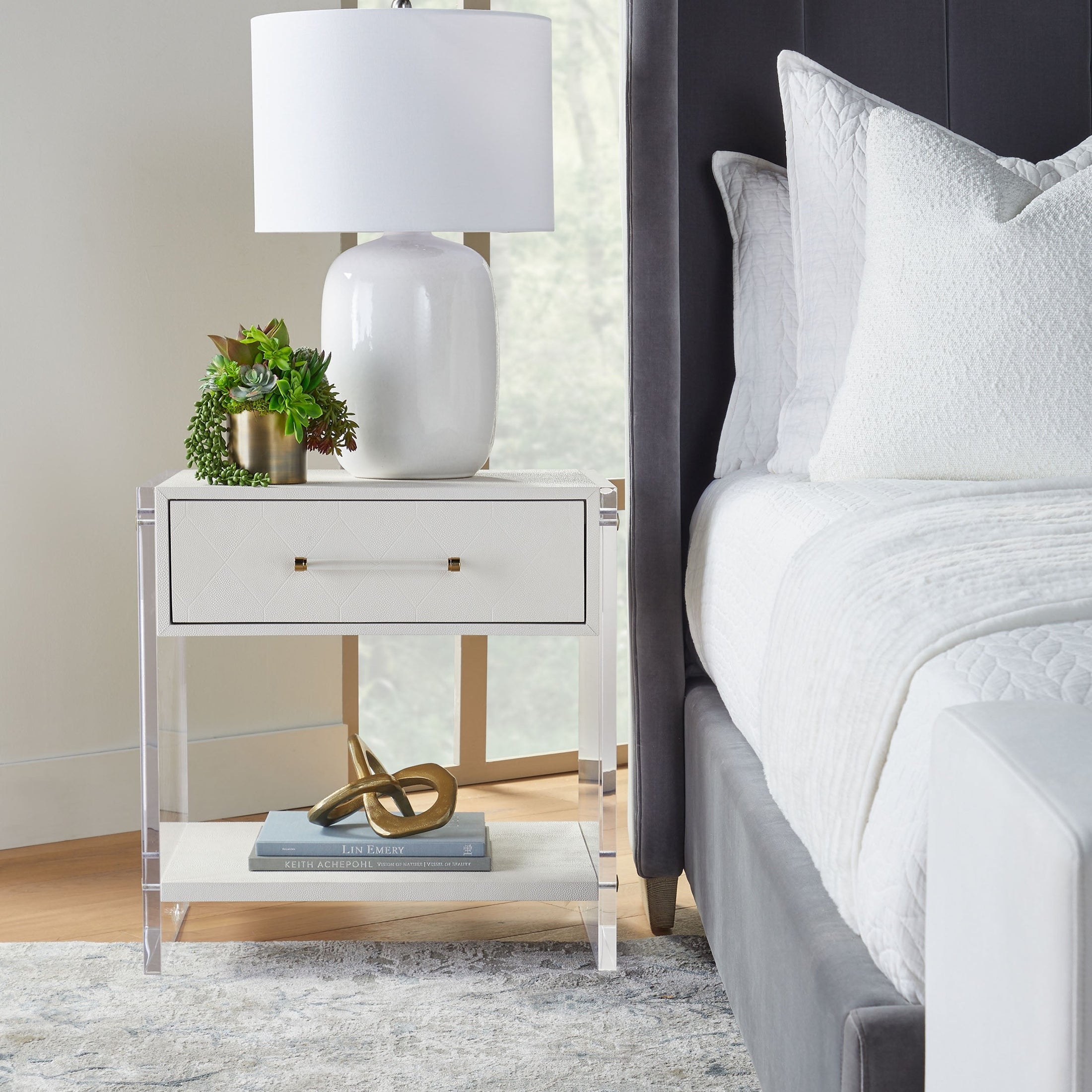 Pearl-white shagreen nightstand with clear acrylic sides and brass hardware beside a gray upholstered bed in a modern bedroom setting