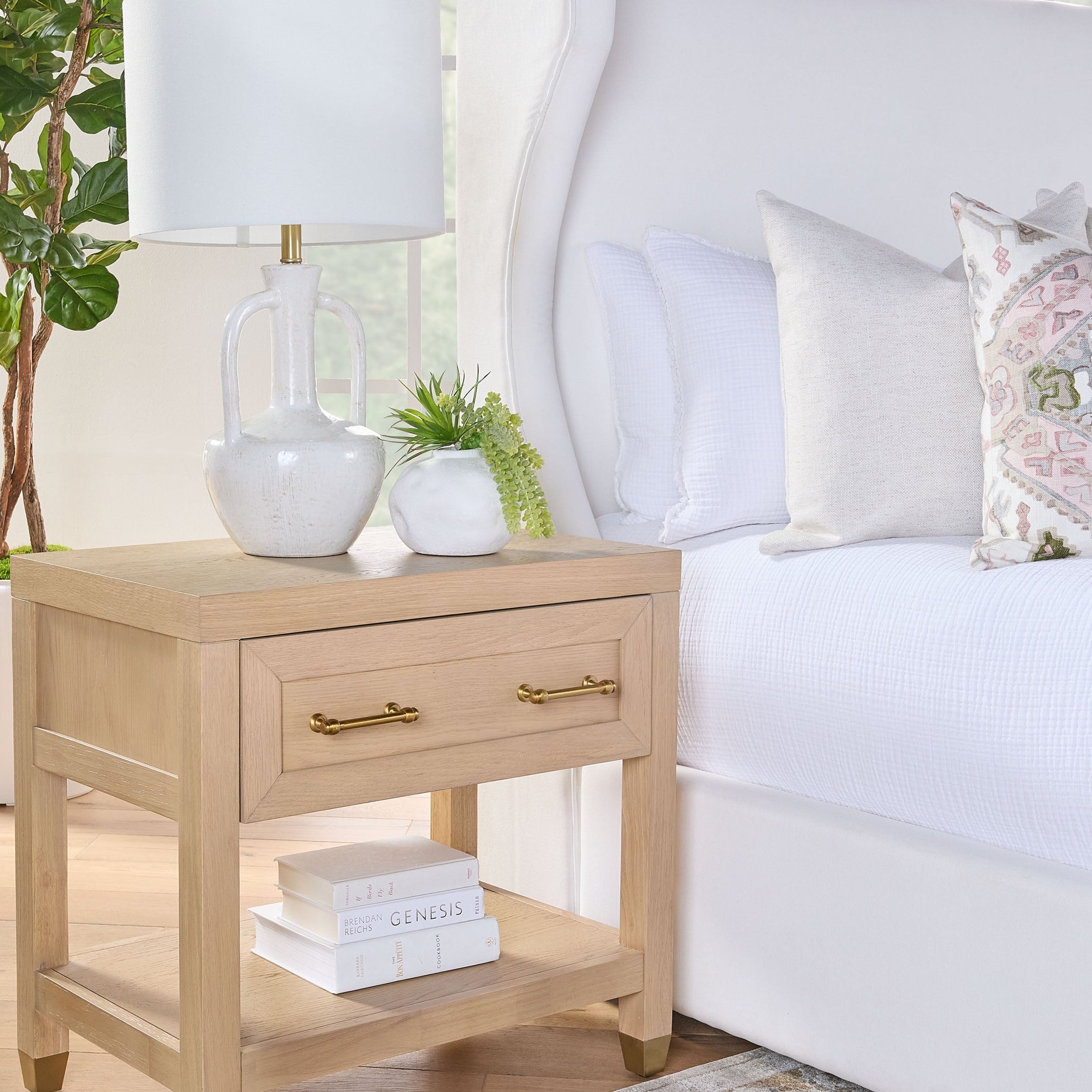 Essentials for Living Stella nightstand in natural wood with brass handles, drawer, lower shelf, and decorative lamp and plant
