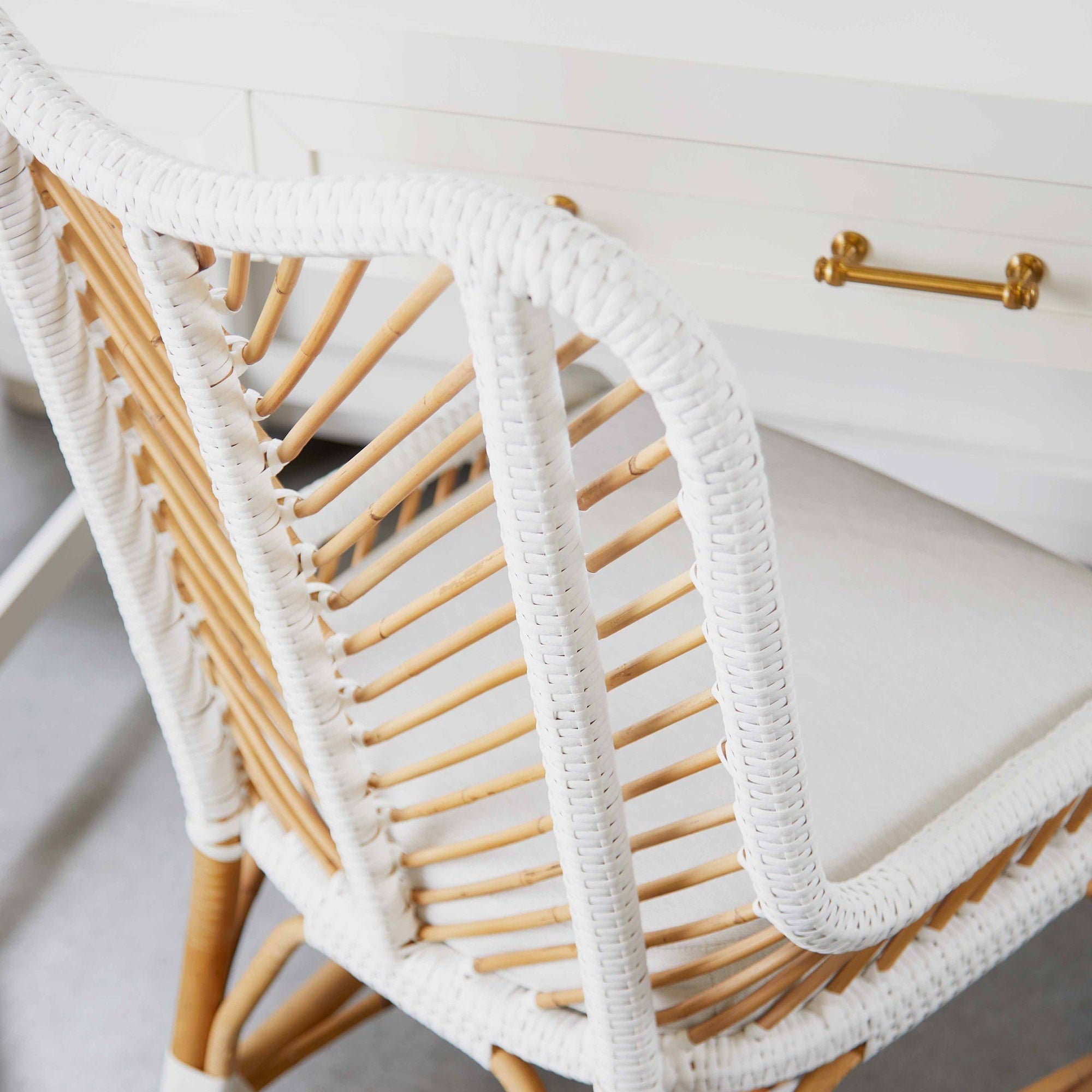Close-up of a white and natural wood rattan chair in front of a white Stella Desk with brass handles and accents