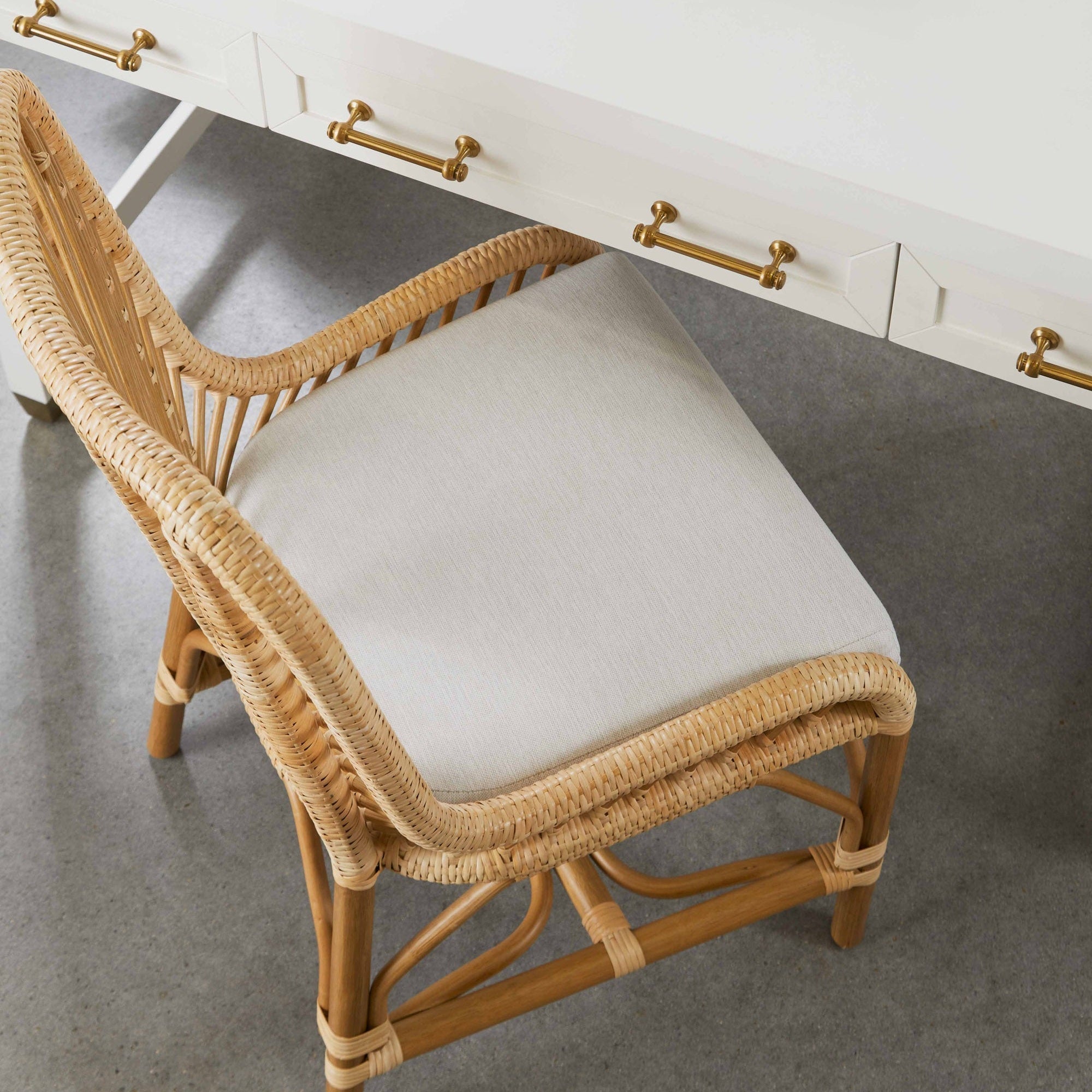Close-up of a white lacquer Stella Desk with brass handles paired with a woven rattan chair with a light cushion.