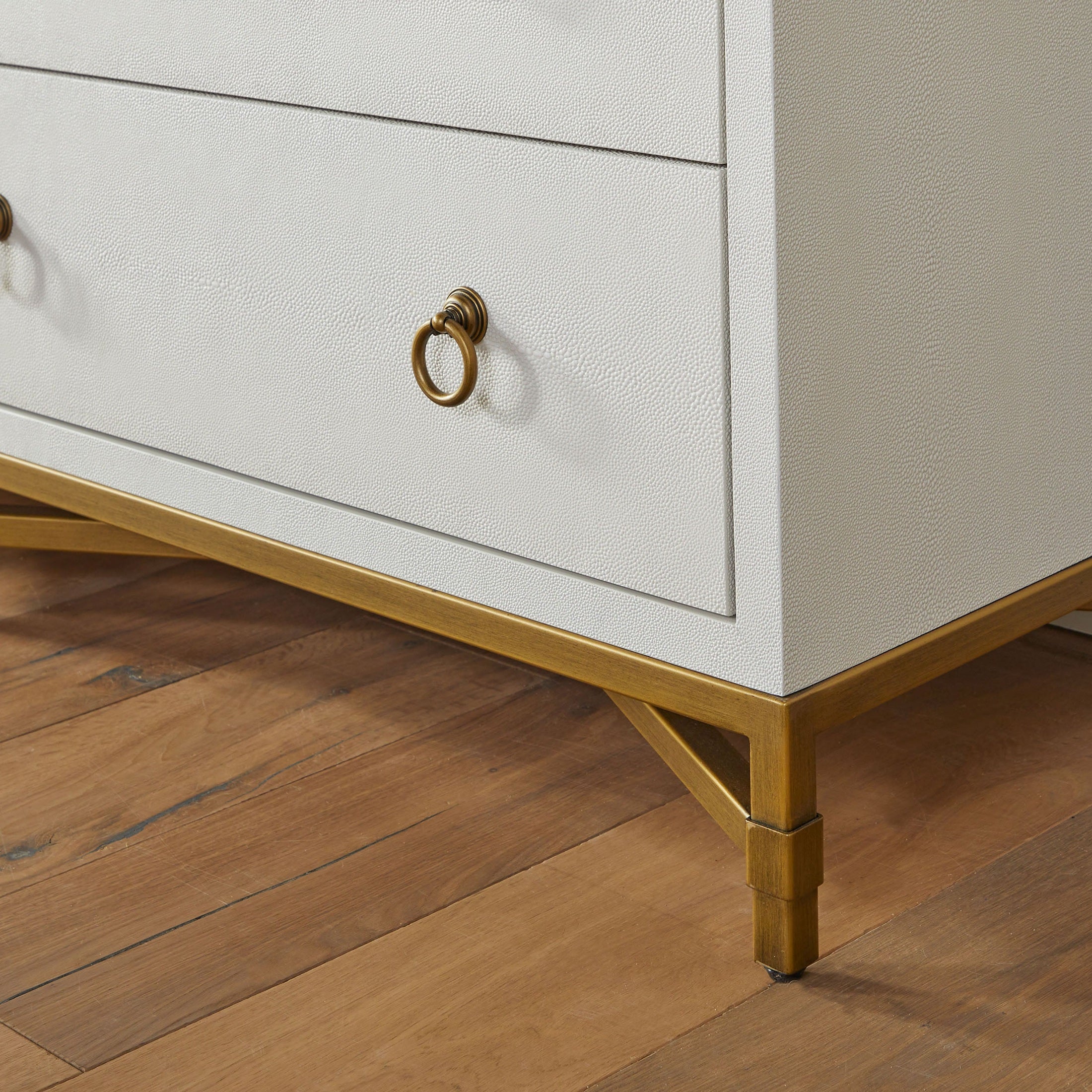 Close-up of pearl white faux shagreen dresser corner with brushed gold ring pulls and X-shaped steel base on wood floor