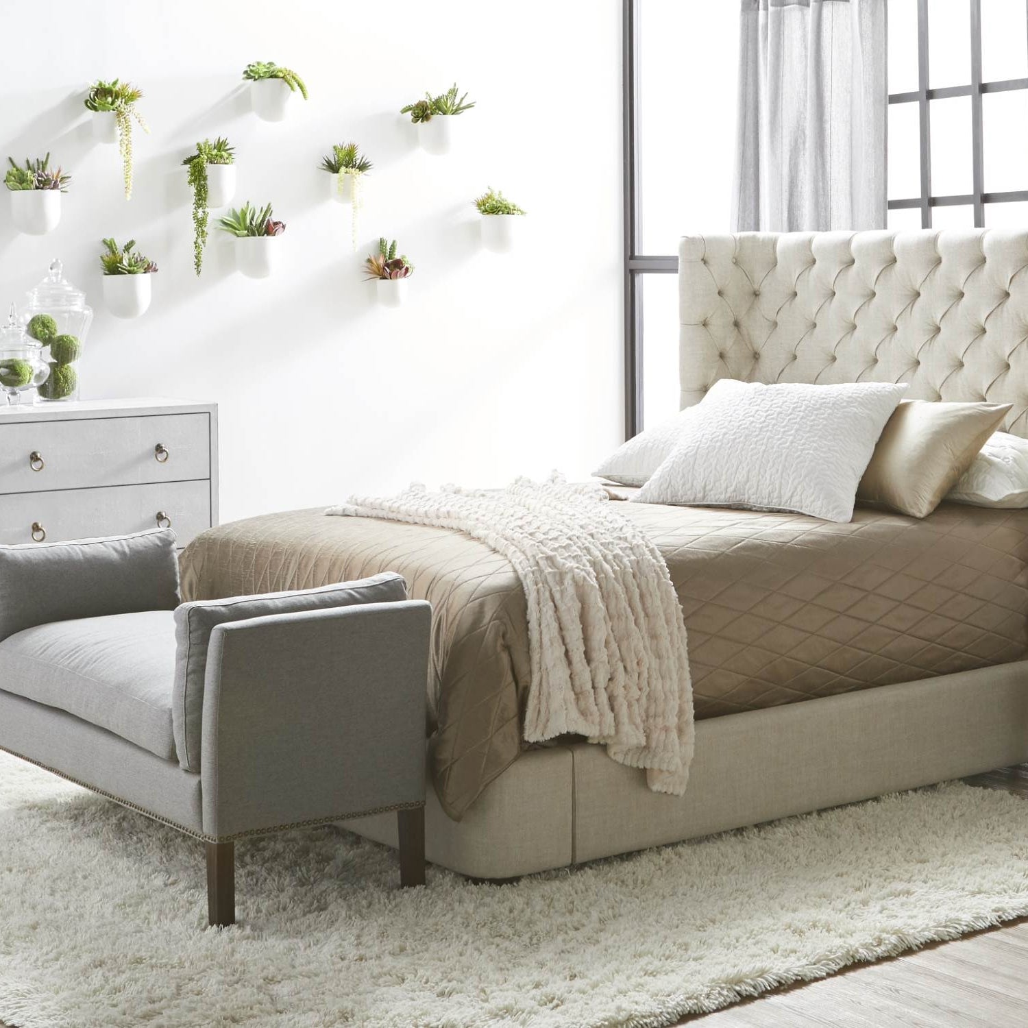 Bright bedroom interior with a tufted bed, beige bedding, gray bench, and floating wall planters in natural light