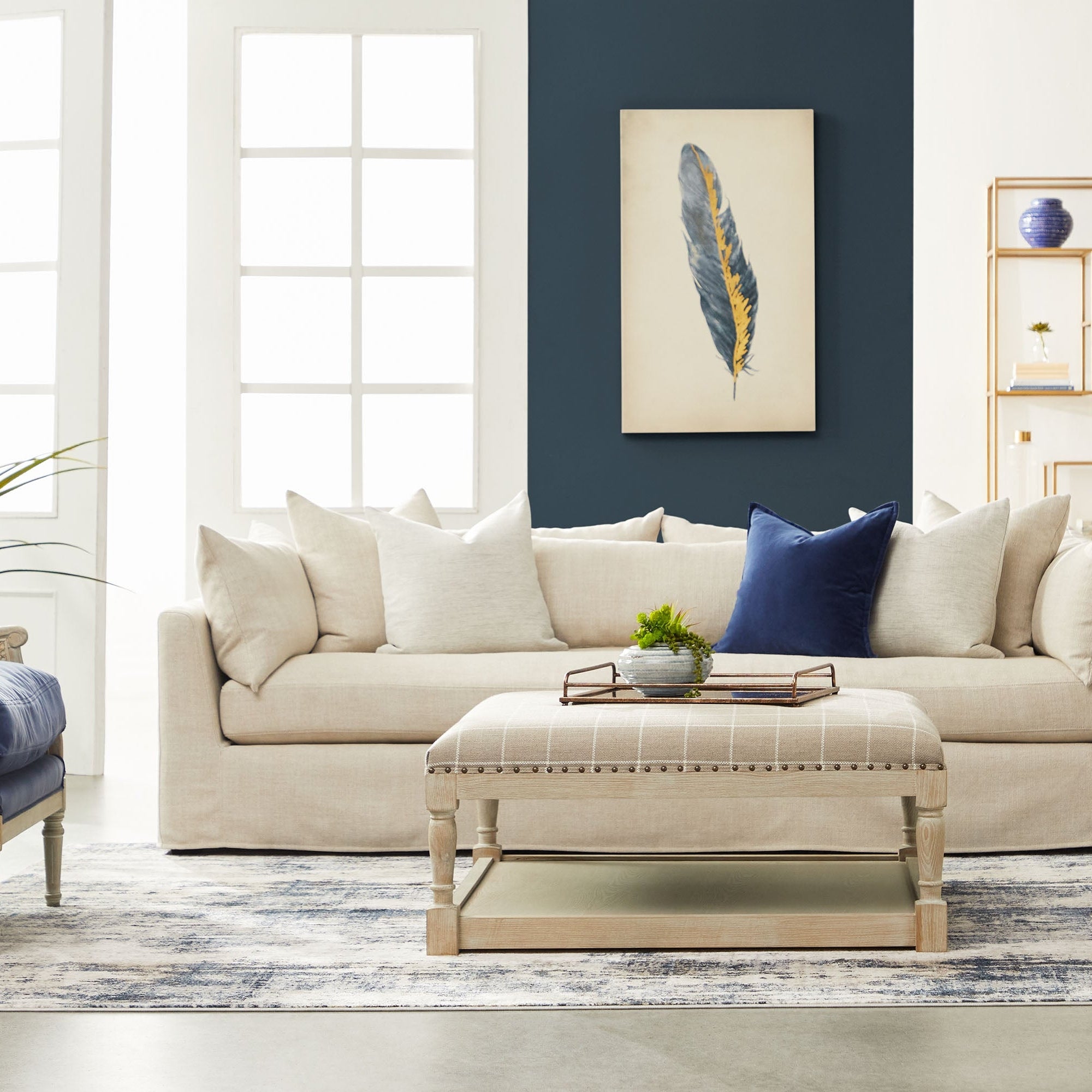 Neutral-toned living room with cream sofa, blue accent pillow, beige ottoman, and dark blue feather wall art