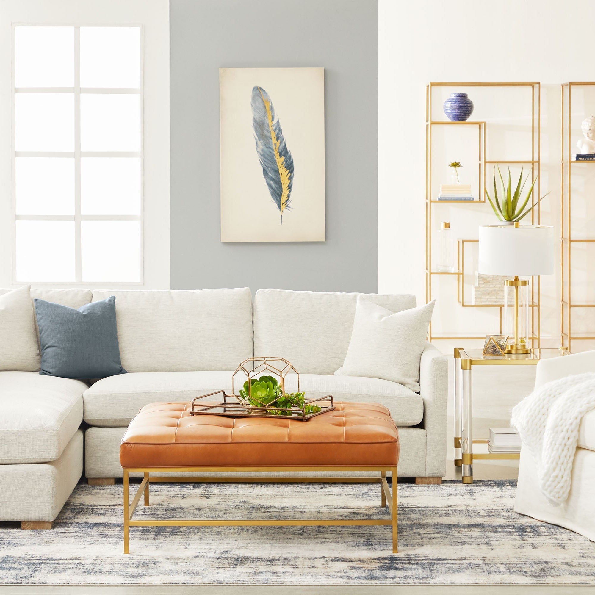 Modern living room with neutral sectional sofa and leather tufted ottoman on patterned rug under natural light