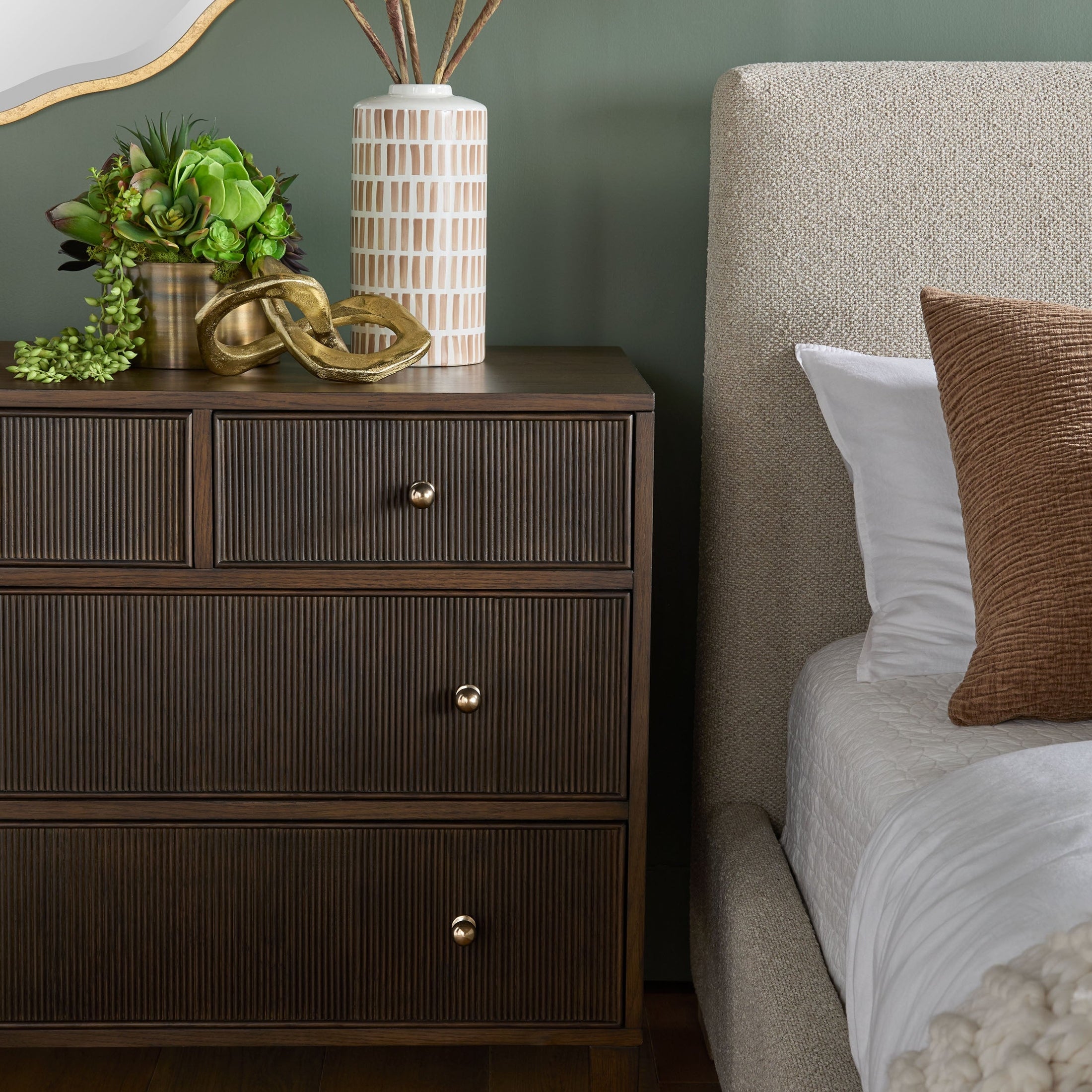 Dark wood dresser with fluted drawer fronts and brass knobs next to upholstered bed with pillows in a modern bedroom setting