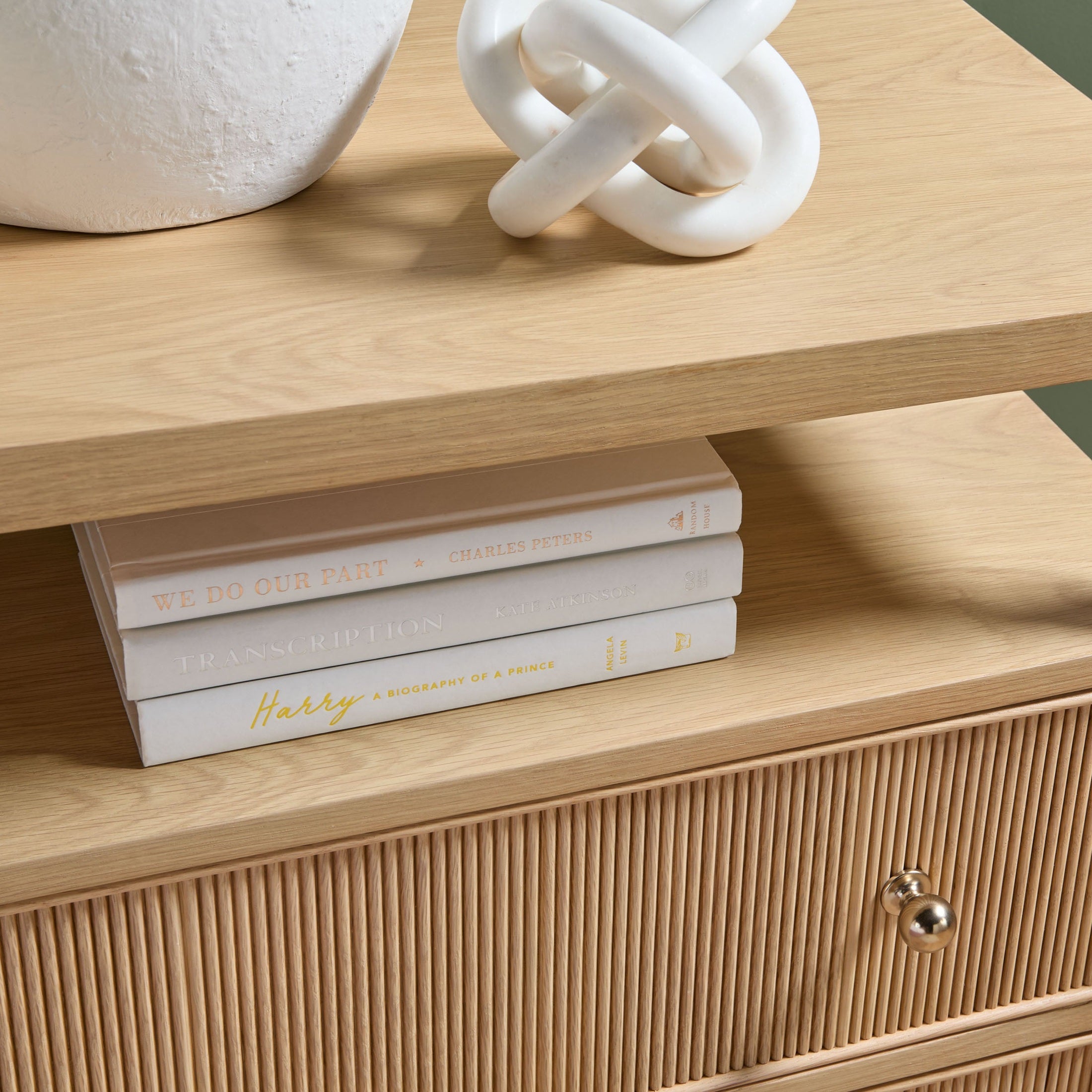 Close-up of oak nightstand with fluted drawer and brass knob, featuring open shelf with stacked books and decor piece