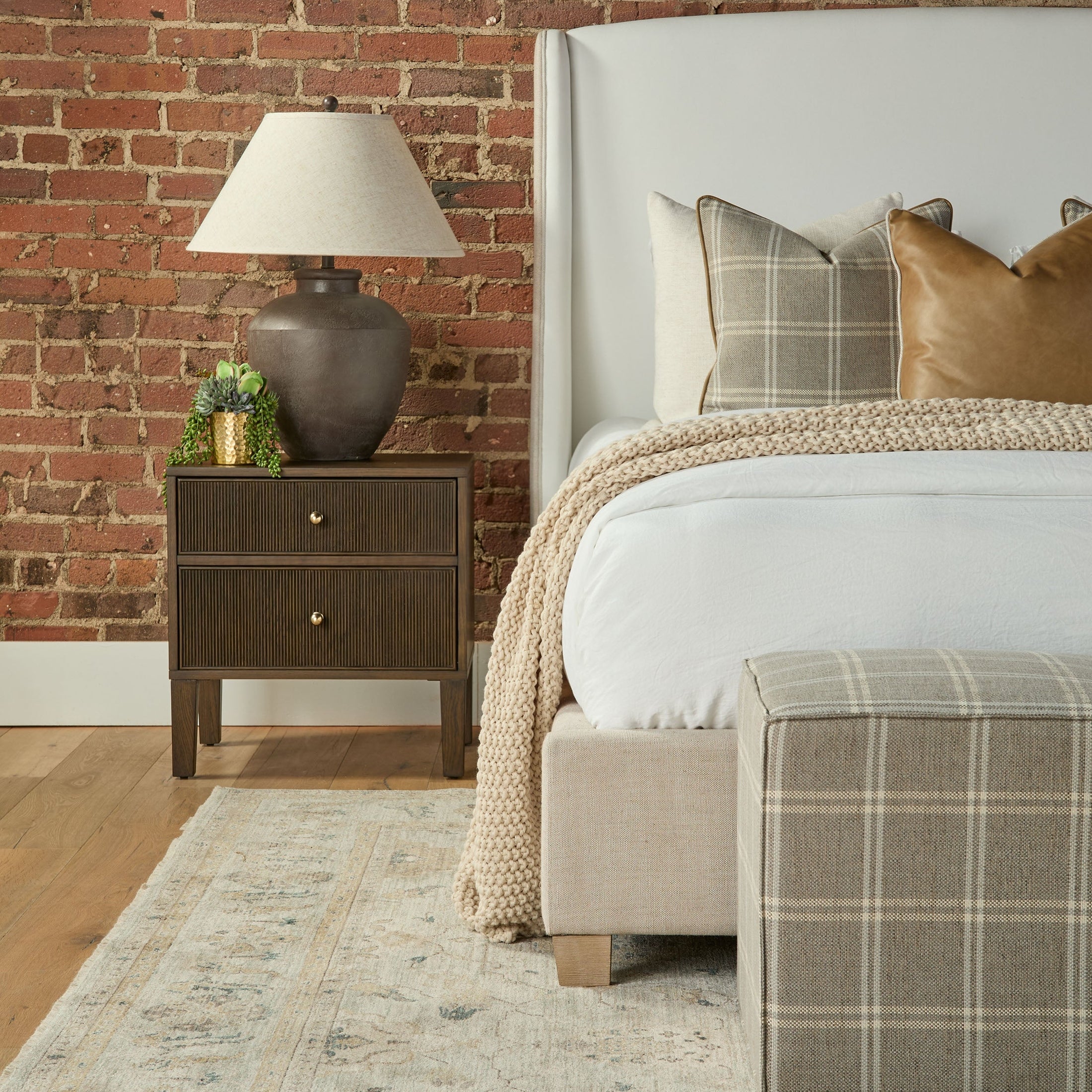 Knox Accent Cube in plaid fabric at the foot of a bed beside a wooden nightstand with lamp and decor