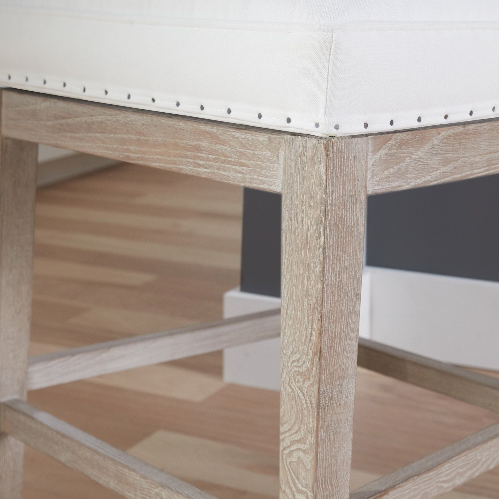Close-up of a Milton Swivel Counter Stool corner showing ash-wood frame in natural gray and Bisque French Linen upholstery with nail-tack trim