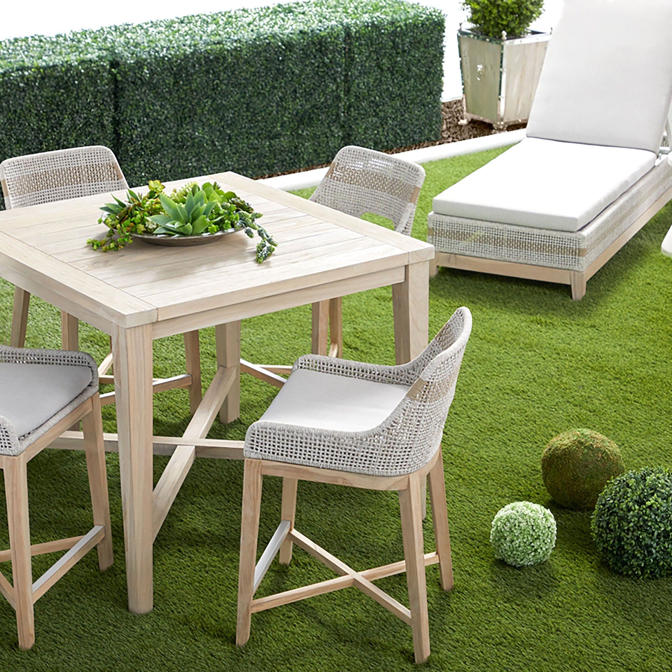 Modern outdoor seating area with woven chairs, square wooden table, chaise lounge, and decorative greenery on grass