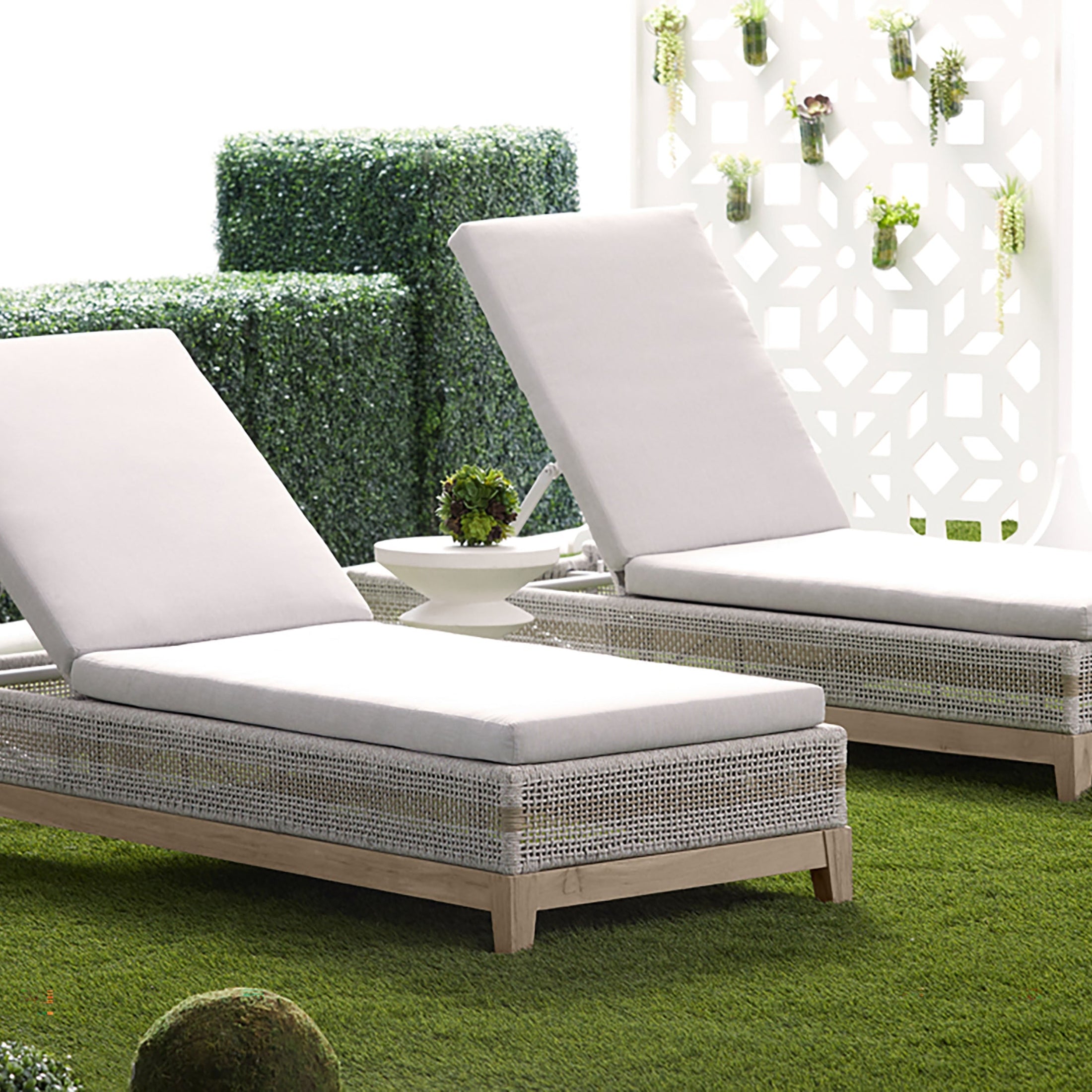 Outdoor lounge chairs with white cushions on green artificial grass near decorative white partition and greenery