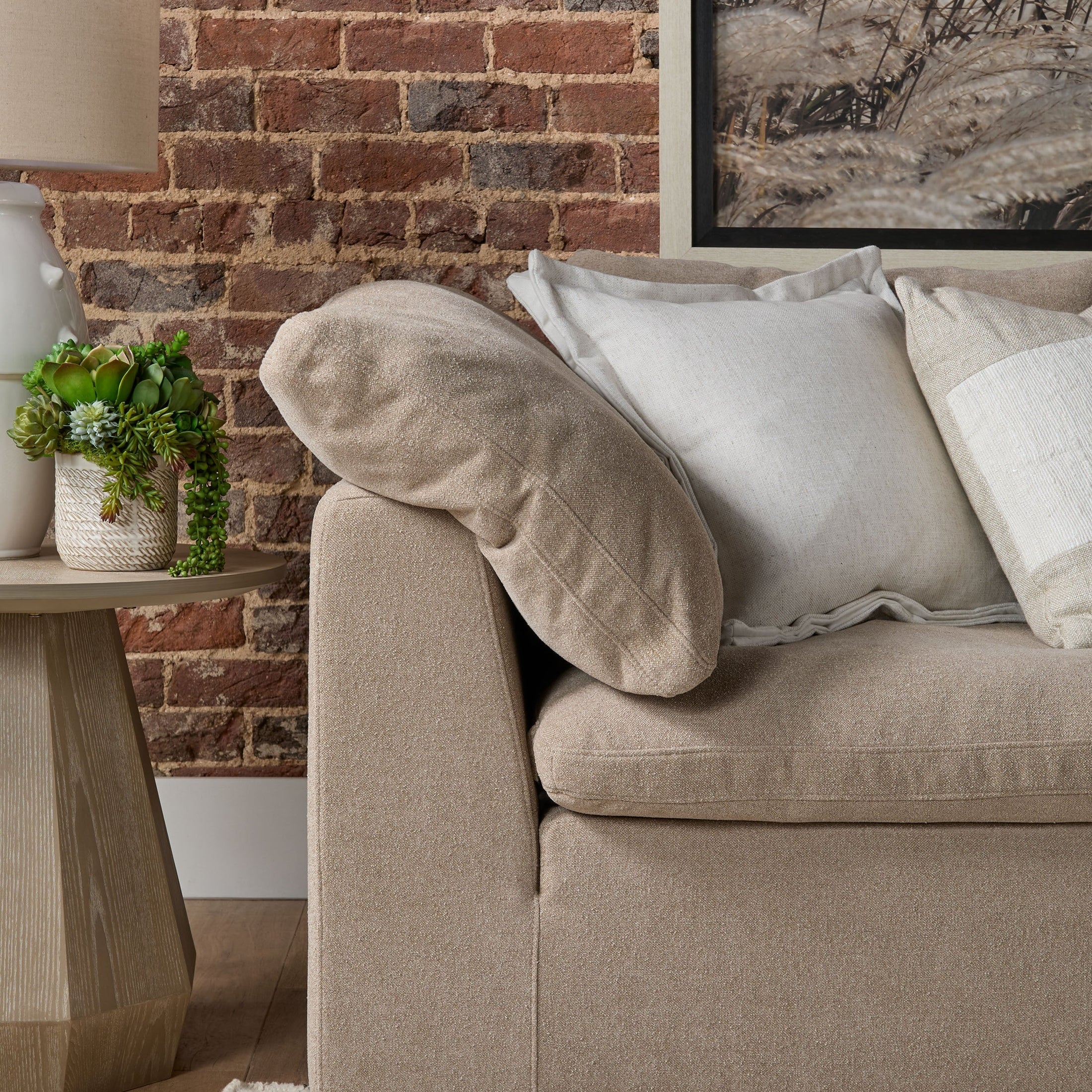 Close-up of beige modular corner chair arm with plush cushions beside wooden side table and brick wall background