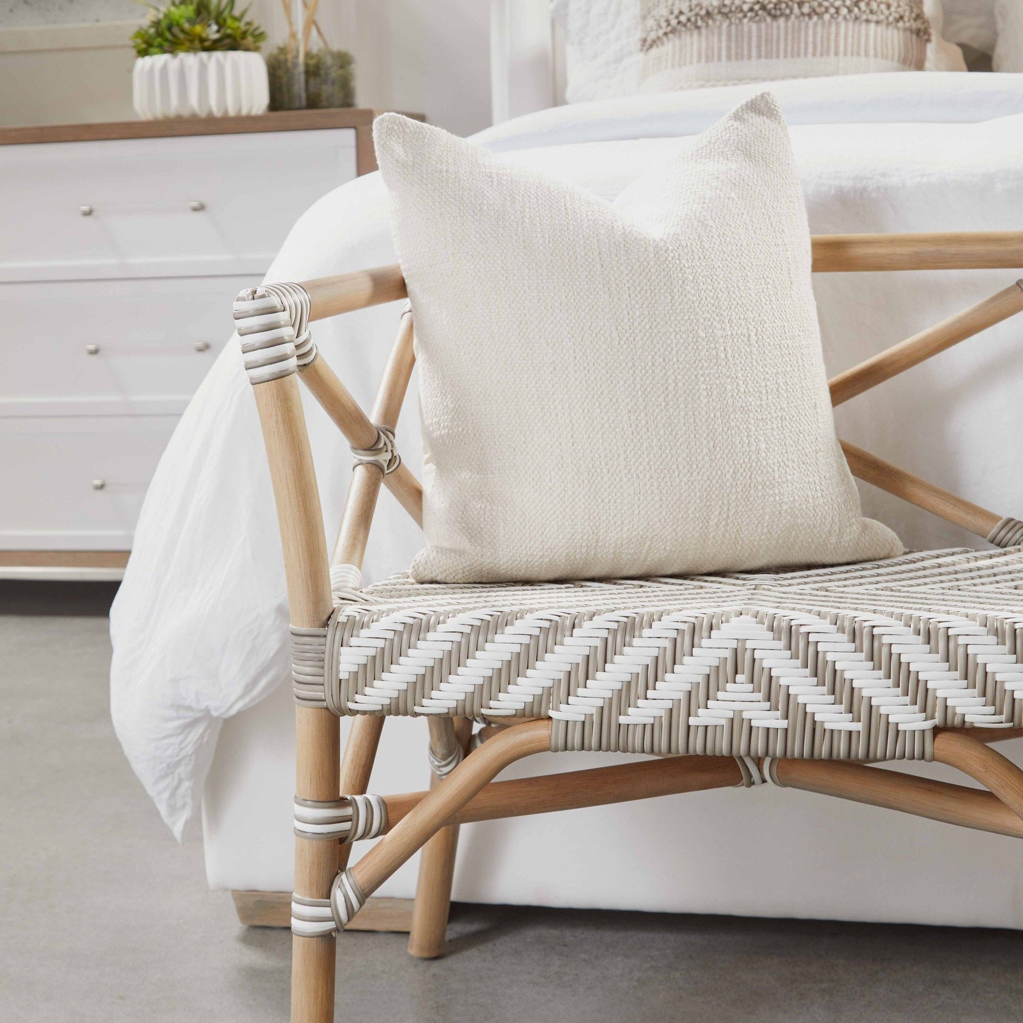 Natural gray oak bench with woven seat and cream textured pillow in bedroom setting with white bedding