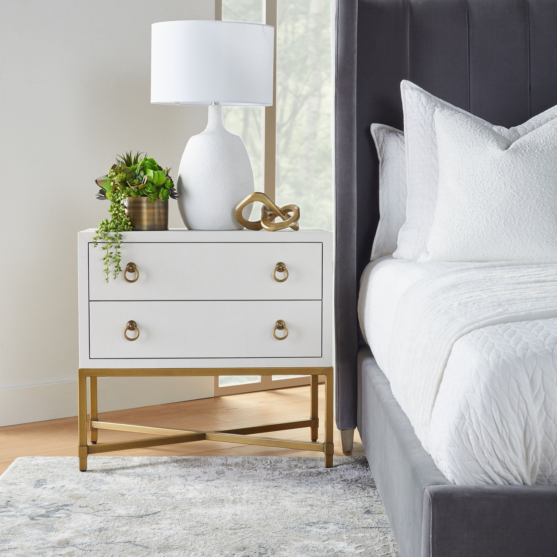 Essentials For Living nightstand with two drawers, white finish, and brushed gold base beside upholstered gray bed