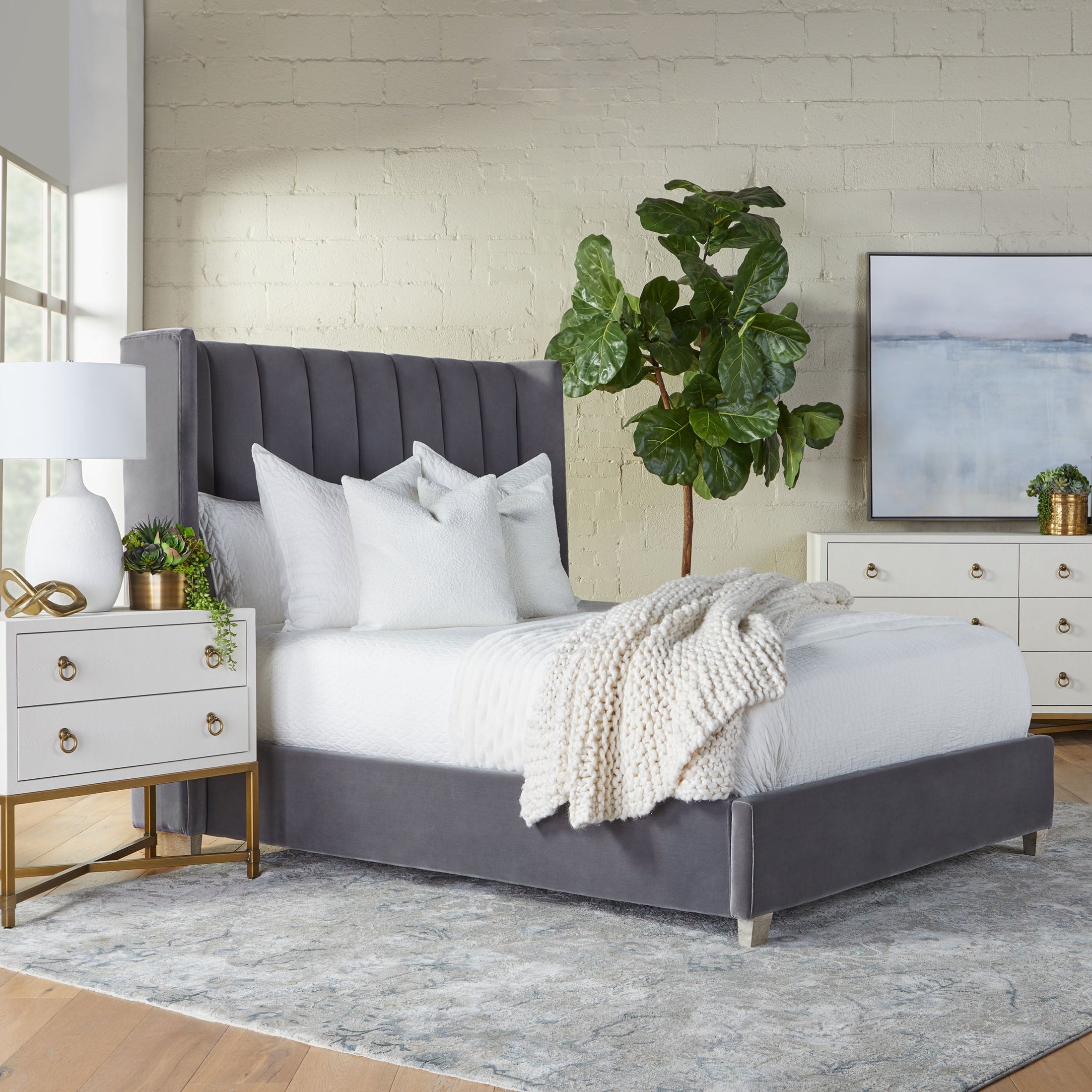 Modern bedroom with gray upholstered bed, white bedding, cream knit throw, and white dresser with gold accents