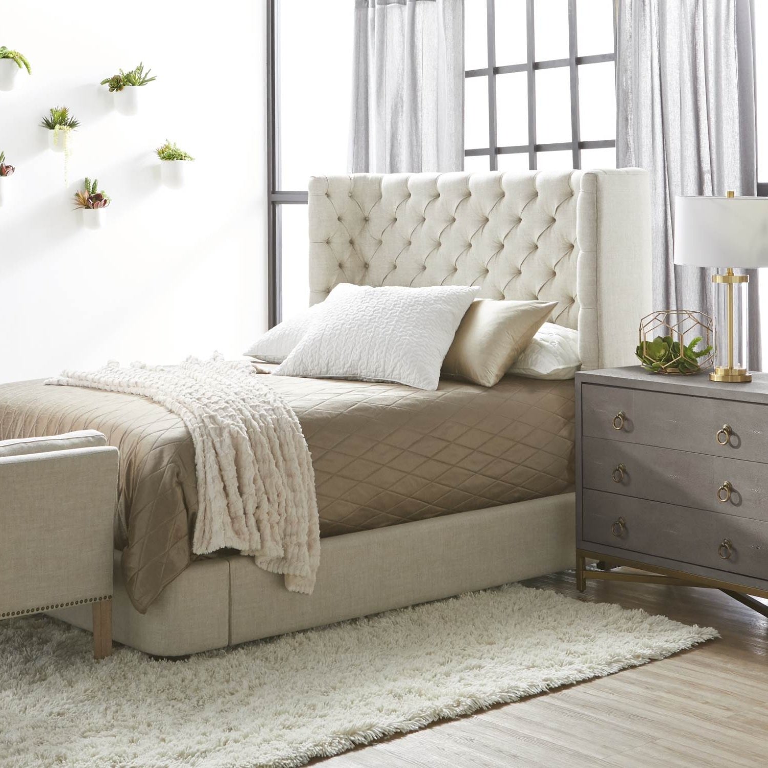 Modern bedroom interior with beige tufted bed, soft bedding, gray dresser, and wall-mounted plants by bright window