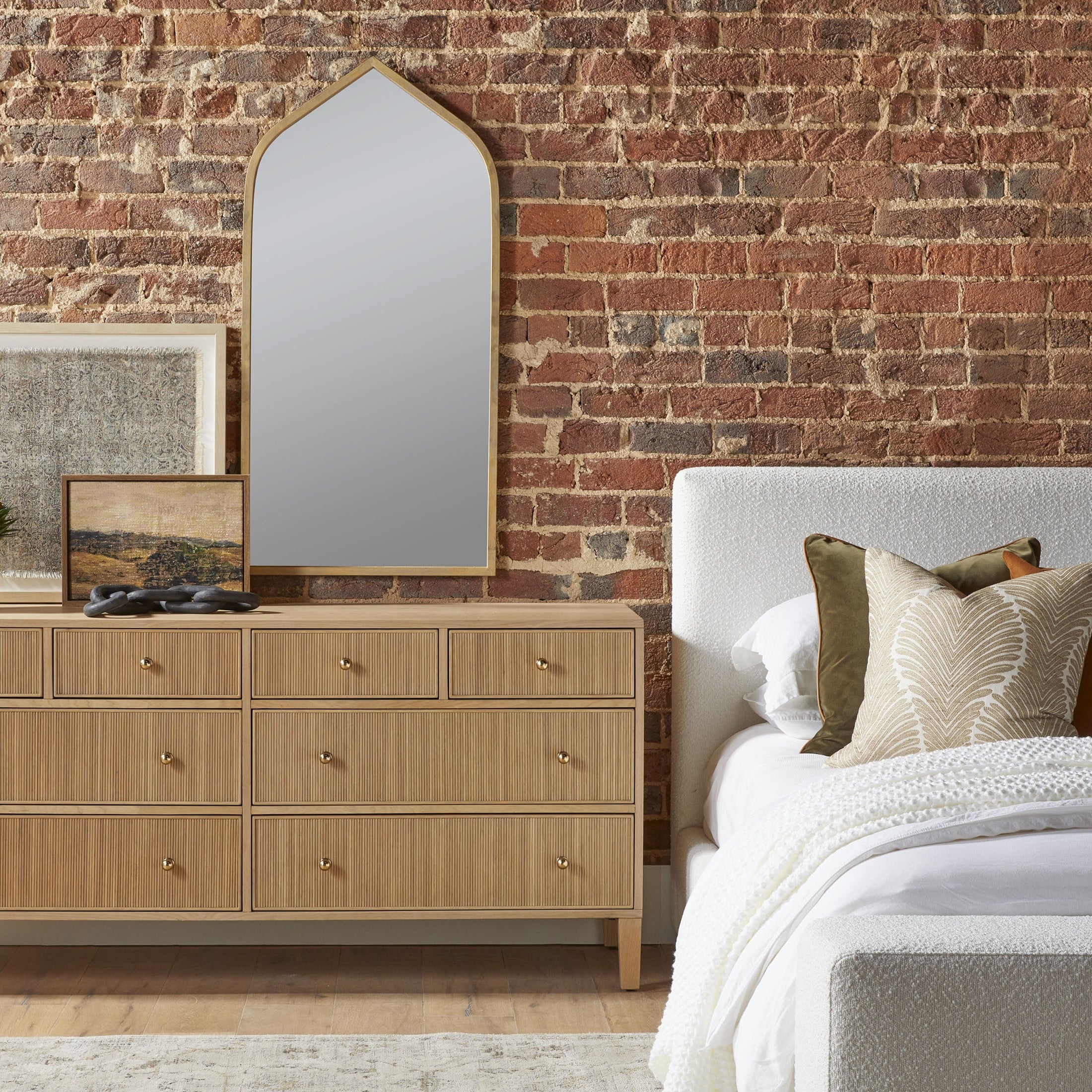 Side view of Warren Standard King Bed upholstered in white fabric with pillows beside wooden dresser and brick wall background